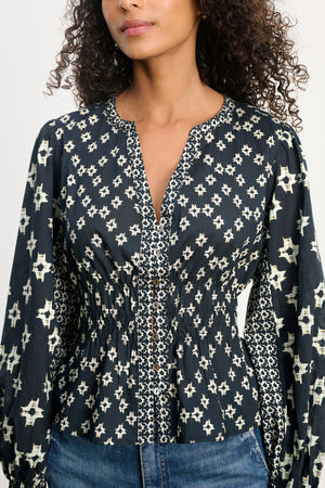 A woman wears the CHARLOTTE TOP by Velvet by Graham & Spencer—a navy blouse with white star prints, breathable cotton, smocked cuffs, V-neckline, button-down front, cinched waist, and long sleeves—paired with blue jeans.