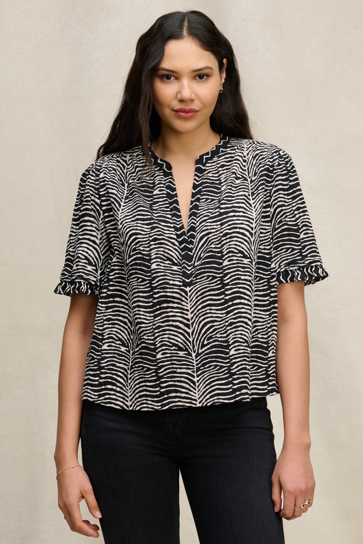 A woman wears the FREEDA TOP by Velvet by Graham & Spencer, a black and white zebra print cotton top with short ruffled sleeves and a V-neckline, paired with black jeans against a neutral background.