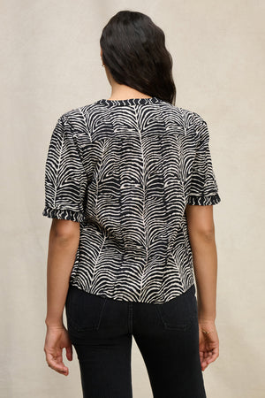 A person shown from the back wears the FREEDA TOP by Velvet by Graham & Spencer—a black and white zebra-striped cotton V-neck top—paired with black jeans against a neutral background.