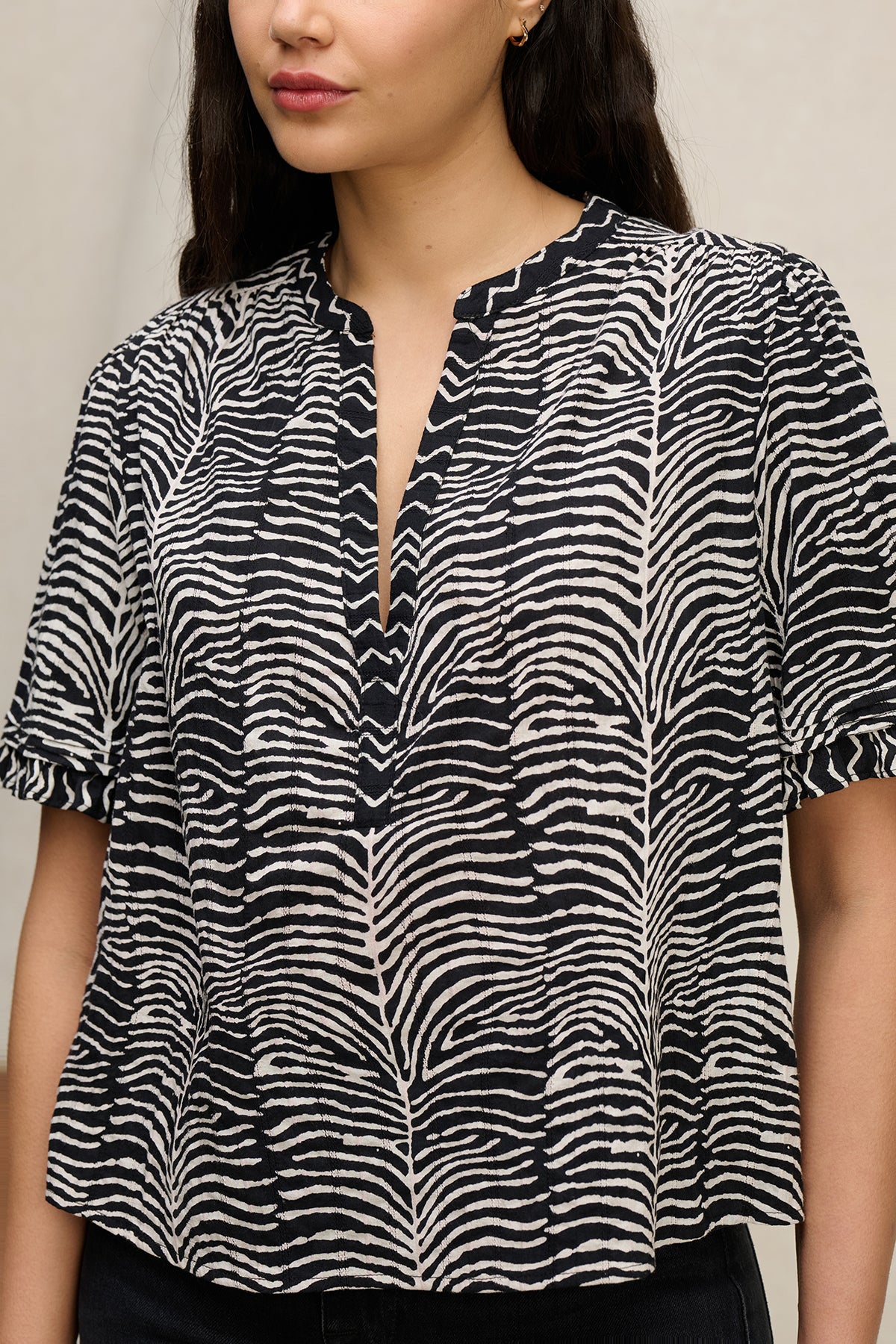 A woman wears the FREEDA TOP by Velvet by Graham & Spencer—a short-sleeved, black and white zebra print cotton top featuring ruffle detailing and a split neckline.