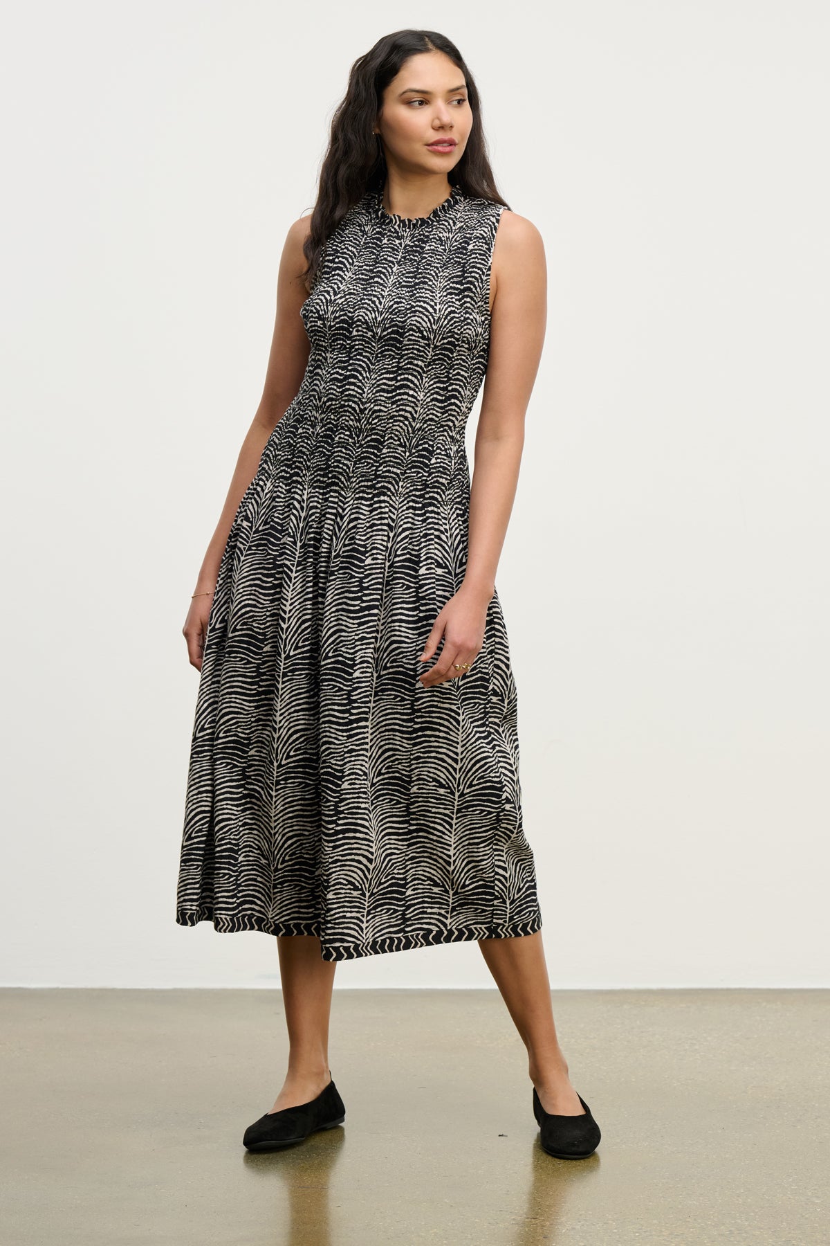 A woman stands against a plain light background wearing the KATHERINE DRESS by Velvet by Graham & Spencer—a sleeveless, black-and-white zebra print cotton midi dress with a smocked bodice, fitted waist, and black flat shoes.