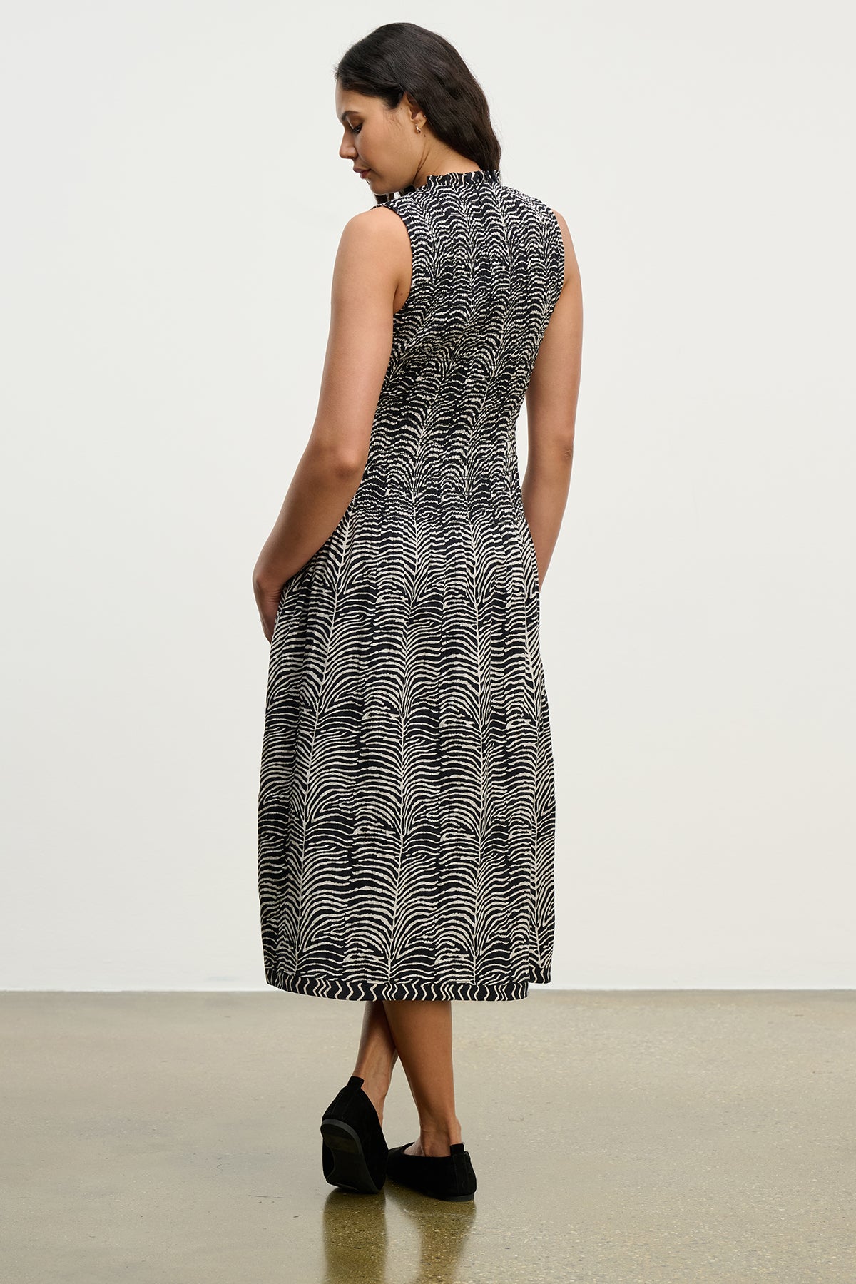 A woman faces away, wearing the Velvet by Graham & Spencer KATHERINE DRESS—a sleeveless zebra print midi with a smocked bodice—and black shoes, set against a plain indoor background.