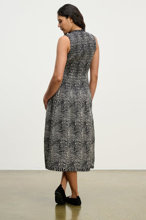 A woman faces away, wearing the Velvet by Graham & Spencer KATHERINE DRESS—a sleeveless zebra print midi with a smocked bodice—and black shoes, set against a plain indoor background.