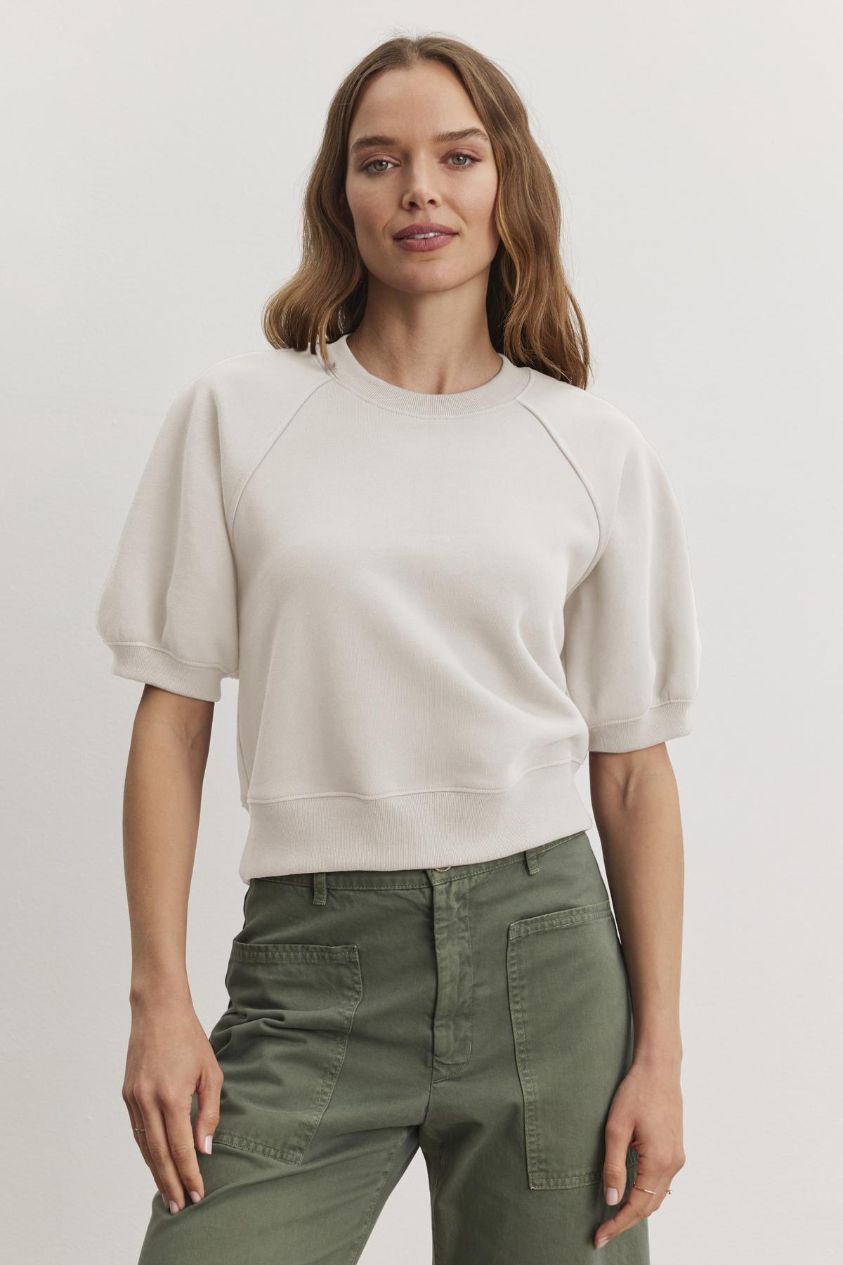 Someone wearing the BERNADETTE SWEATSHIRT by Velvet by Graham & Spencer, a light-colored cropped short-sleeve design in soft cotton fleece, and green pants stands against a plain background.-38512380936385