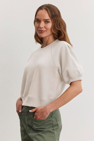 The person with long hair is wearing the BERNADETTE SWEATSHIRT by Velvet by Graham & Spencer, a white short-sleeve cropped sweater, paired with green pants. They are standing sideways with hands in pockets against a plain background.