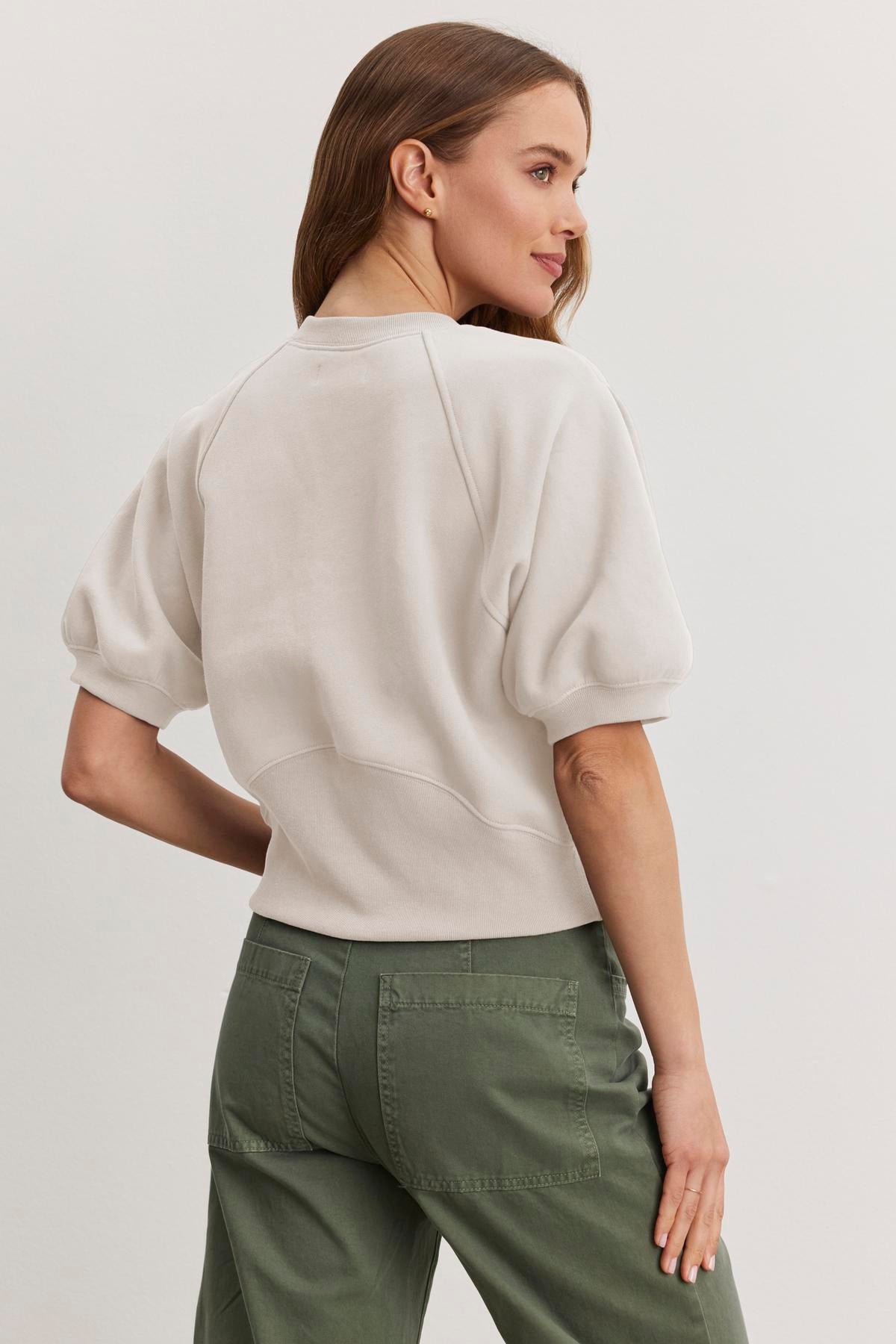 A woman wears the BERNADETTE SWEATSHIRT from Velvet by Graham & Spencer, a cropped design in white cotton fleece, paired with green pants while facing away.-38512381001921
