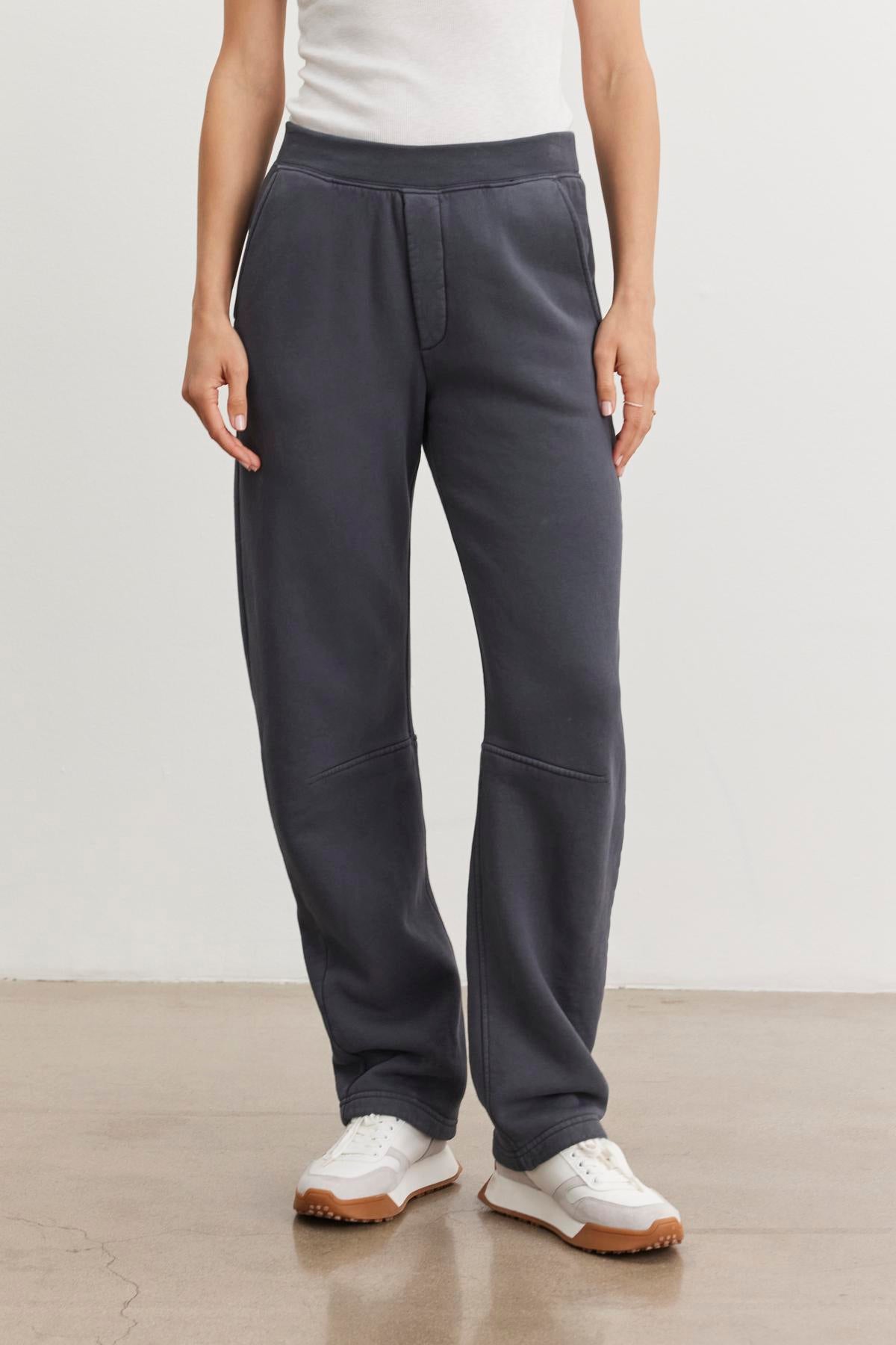 A person dressed in a cozy white shirt and wearing the MATTI SWEATPANT by Velvet by Graham & Spencer, along with white sneakers, stands on a concrete floor against a white background.
