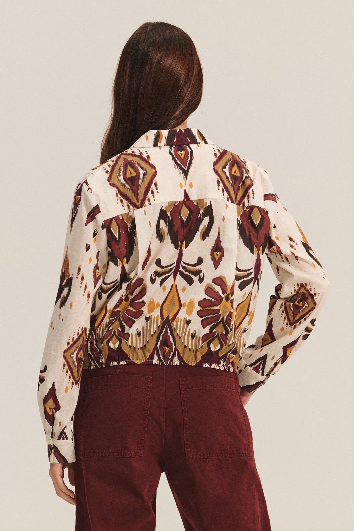 A person with long brown hair is shown from the back wearing the KAYLA TOP by Velvet by Graham & Spencer, a cotton-viscose blend button-down featuring geometric and abstract designs, paired with burgundy pants.-40681722839233
