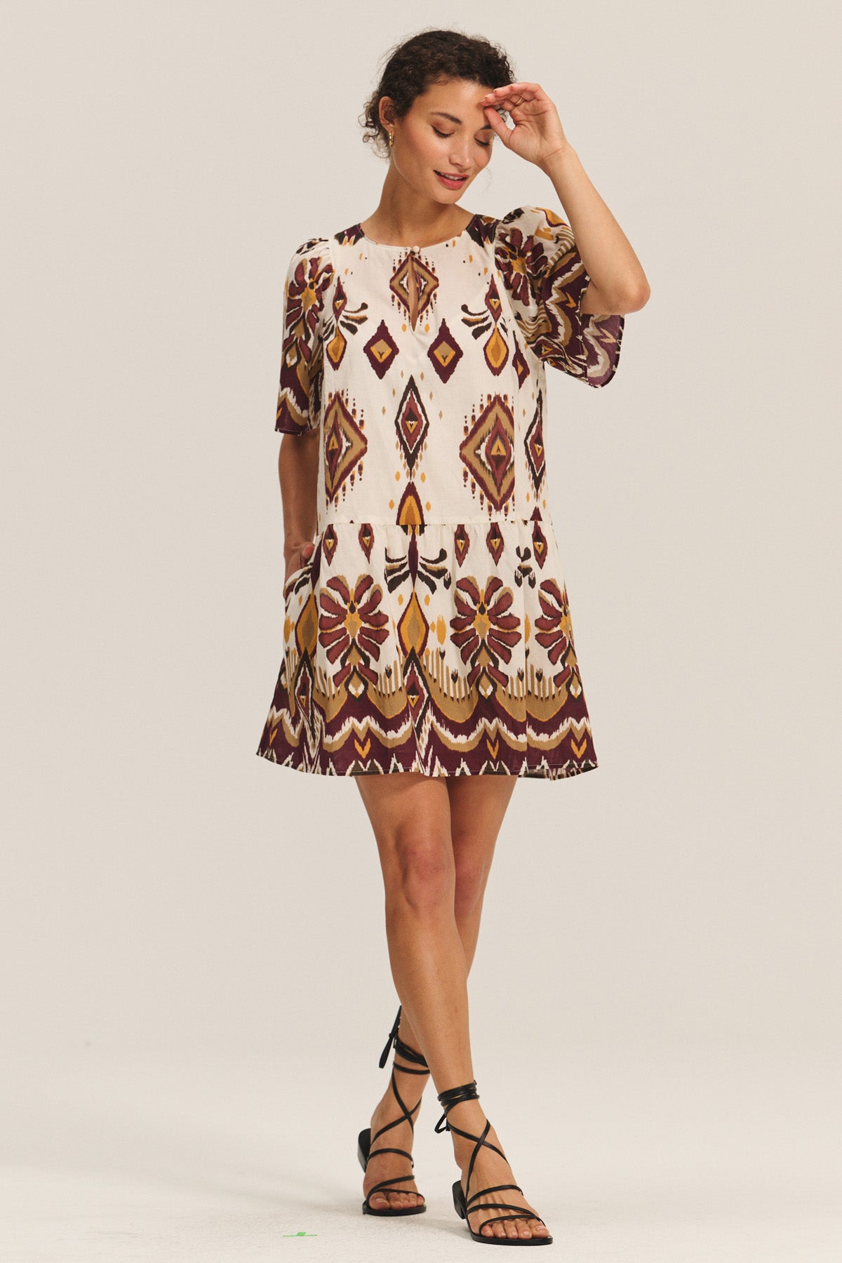 A woman stands in a relaxed pose wearing the MAGGIE DRESS by Velvet by Graham & Spencer, featuring geometric and floral prints in earthy tones, paired with black strappy sandals and finished with an adjustable belt.-40598485565633