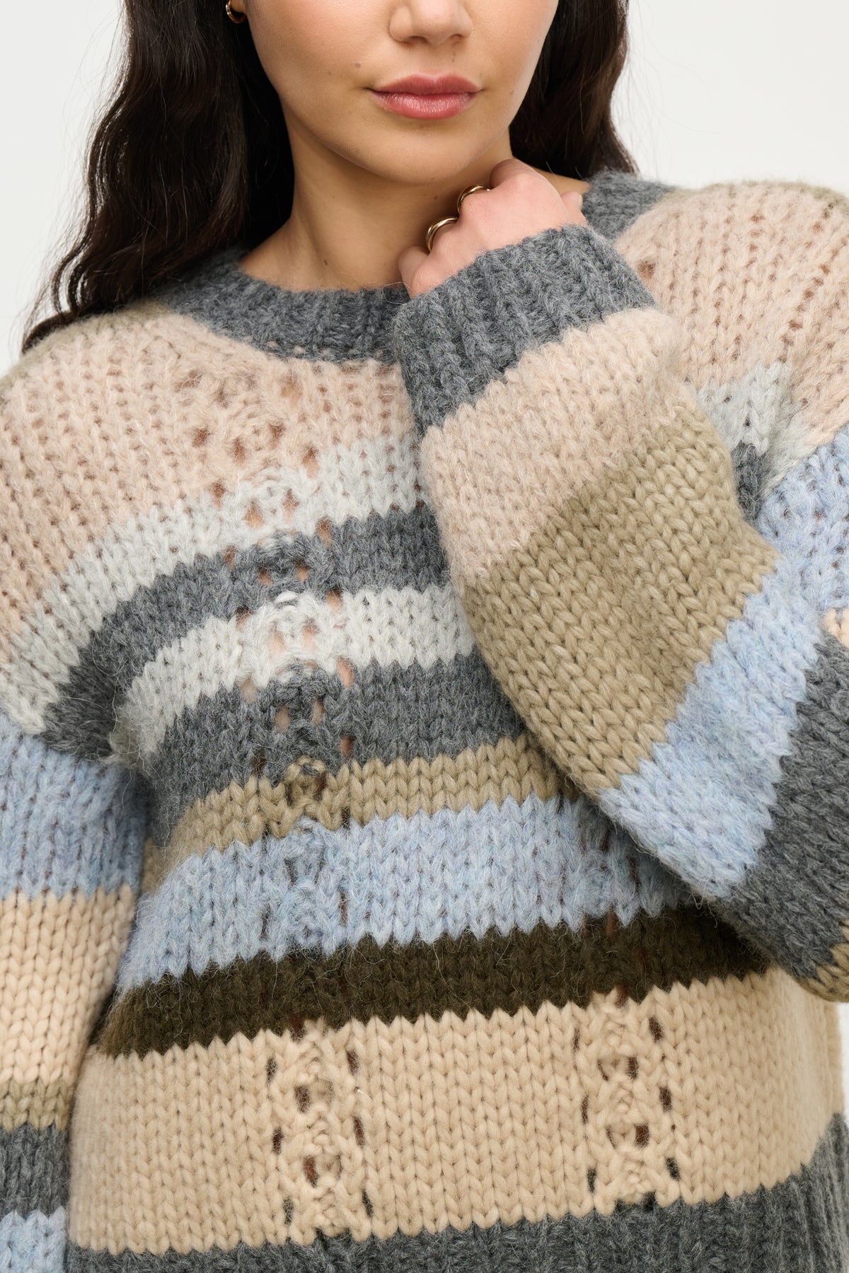 A woman wears the RINA ALPACA SWEATER by Velvet by Graham & Spencer—a boxy, chunky knit with wide horizontal stripes in beige, blue, gray, and olive green. She touches her face with her left hand.-Hover Image