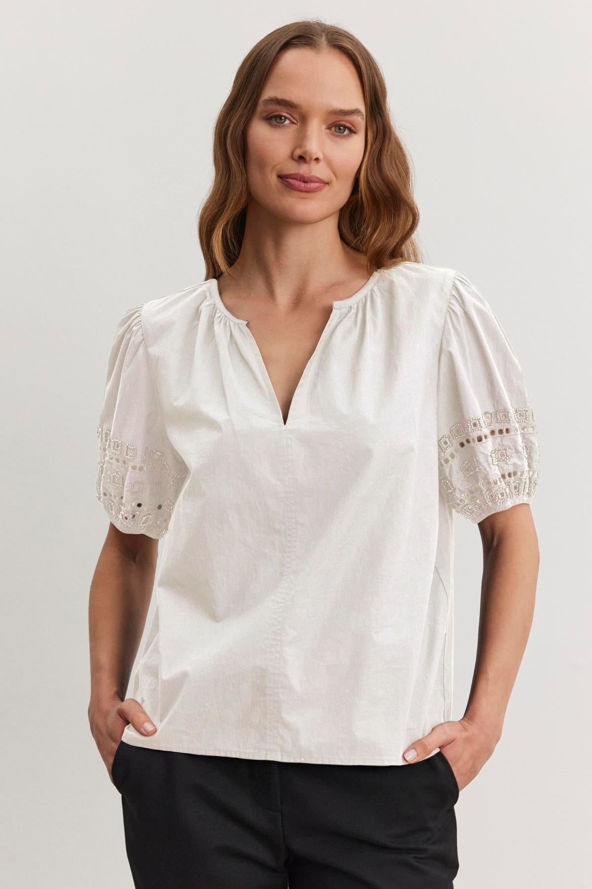 A person in the MARTAH TOP by Velvet by Graham & Spencer, featuring a white cotton poplin fabric with lace puff sleeves, and black pants stands with hands in pockets against a plain background.