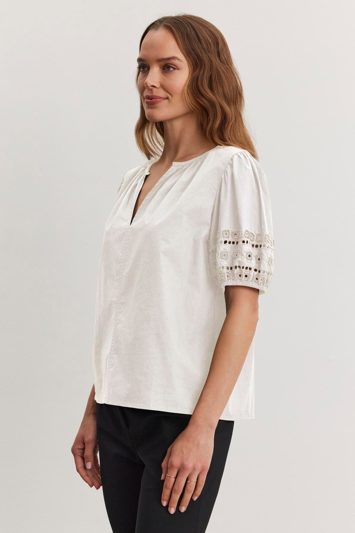 A woman with long hair wears the MARTAH TOP from Velvet by Graham & Spencer, characterized by a v-neckline and lace-detailed puff sleeves. She pairs it with black pants, standing against a plain background.