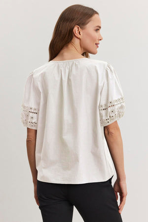 In a plain setting, a person faces away wearing the MARTAH TOP, a white v-neck blouse with lace sleeves by Velvet by Graham & Spencer, paired with black pants.