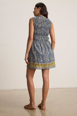 A woman stands indoors facing away, wearing the sleeveless MARLA DRESS by Velvet by Graham & Spencer and sandals, on a light-colored floor against a plain background.