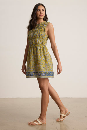 A woman stands indoors in the MARLA DRESS by Velvet by Graham & Spencer, a yellow block-printed cotton mini with blue accents and a ruffle round neckline, paired with beige sandals, facing slightly to the side.