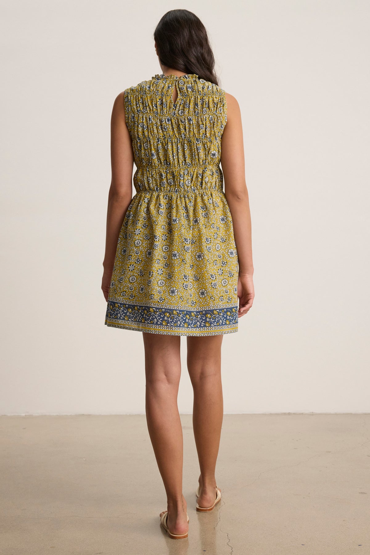 A woman stands facing away, wearing the MARLA DRESS by Velvet by Graham & Spencer—a sleeveless yellow block-printed cotton mini dress with a gathered waist and ruffled neckline—paired with flat sandals on a plain background.