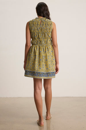 A woman stands facing away, wearing the MARLA DRESS by Velvet by Graham & Spencer—a sleeveless yellow block-printed cotton mini dress with a gathered waist and ruffled neckline—paired with flat sandals on a plain background.
