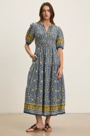 A woman poses against a plain background in the NATALIA DRESS by Velvet by Graham & Spencer—a blue and yellow floral cotton dress with a smocked waist, ankle-length hem, and short puff sleeves, styled with sandals.