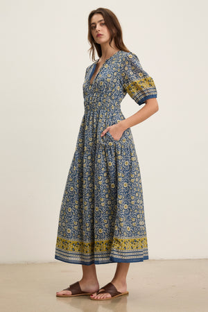 A woman wearing the Velvet by Graham & Spencer NATALIA DRESS, a blue and yellow floral cotton maxi with a smocked waist and short sleeves, paired with brown sandals, stands against a plain light background.