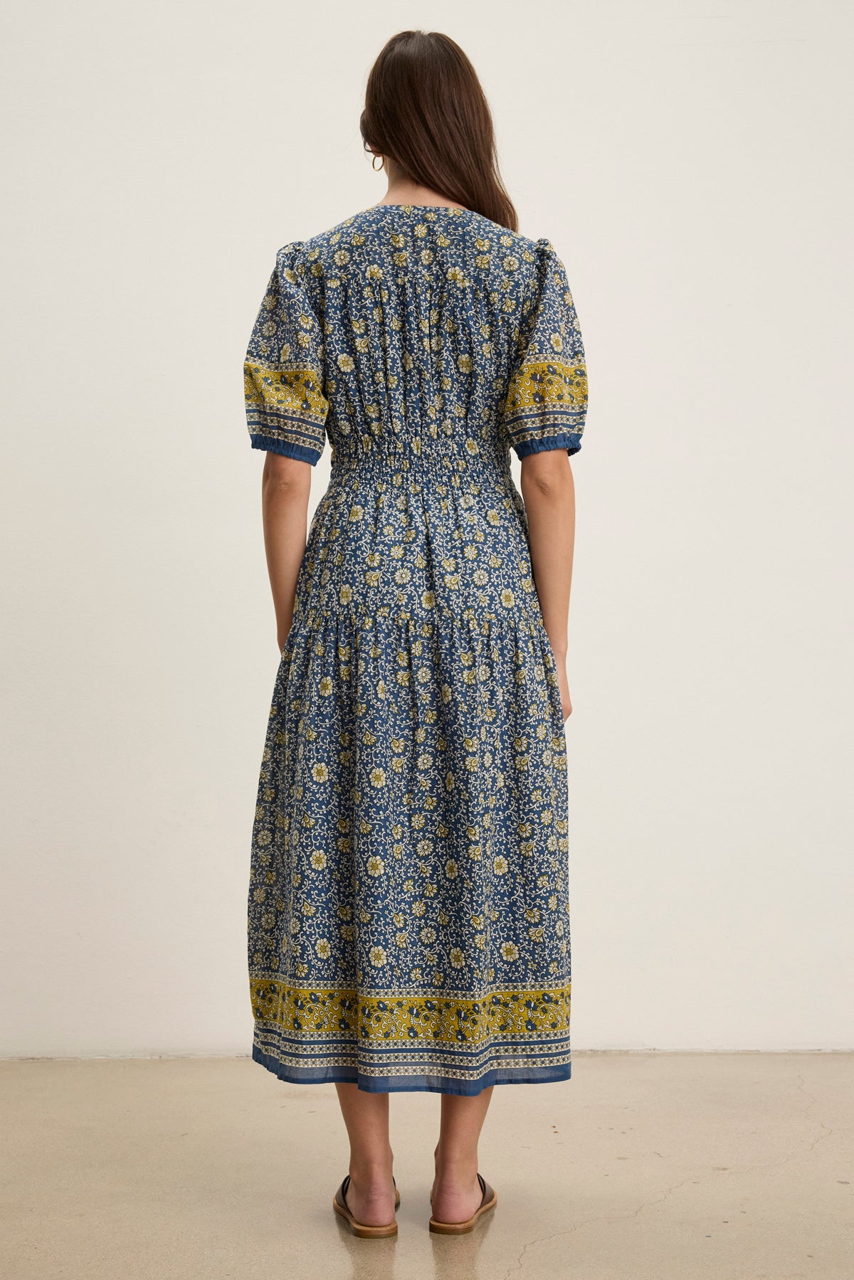 A woman faces away, wearing the NATALIA DRESS by Velvet by Graham & Spencer—a blue and yellow block print, short-sleeved, ankle-length cotton dress with a smocked waist and flat shoes against a plain background.-39120097083585