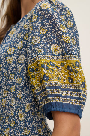 Close-up of a person wearing the NATALIA DRESS by Velvet by Graham & Spencer—a blue printed cotton dress with white and yellow florals, puffed short sleeves accented by a yellow band, and a smocked waist for shape.