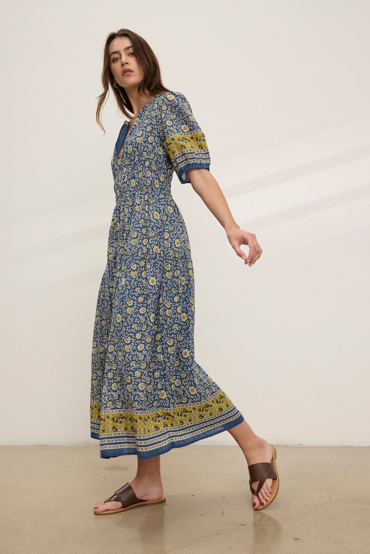A woman wears the NATALIA DRESS by Velvet by Graham & Spencer—a blue and yellow printed cotton dress with a smocked waist—paired with brown sandals, standing against a plain light-colored wall.