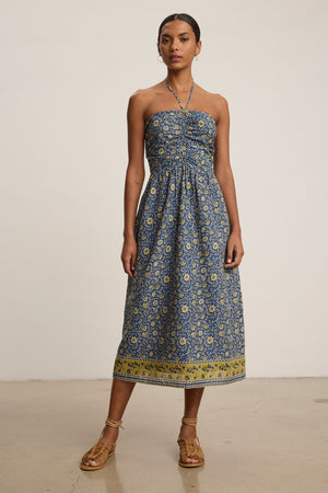 A woman wears the ODETTE DRESS by Velvet by Graham & Spencer, a chic blue and yellow strapless midi floral dress, paired with brown sandals against a plain background.