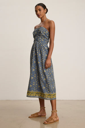 A woman stands indoors in the ODETTE DRESS by Velvet by Graham & Spencer—a chic blue and yellow block print cotton midi dress—paired with sandals, against a plain background.