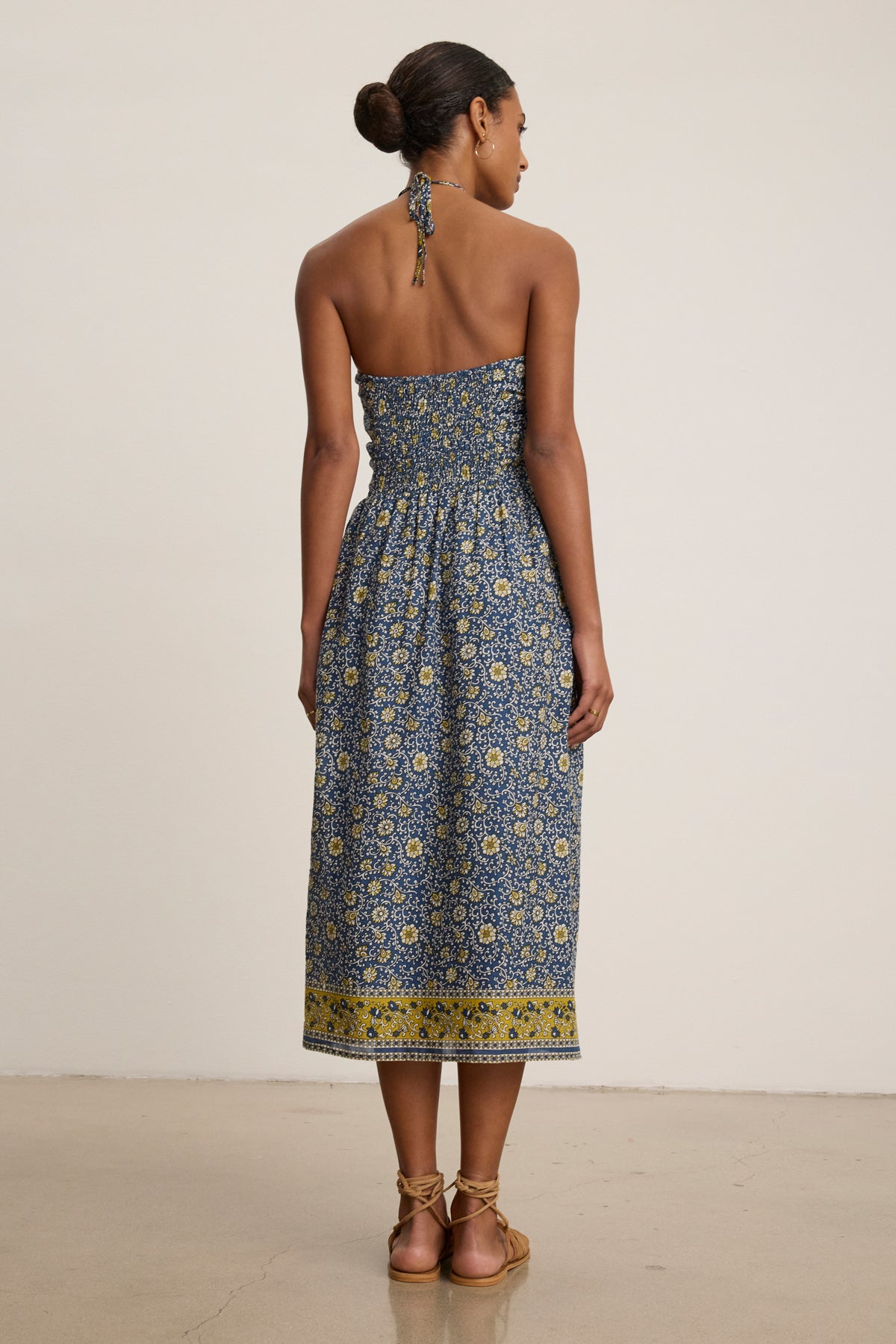 A woman faces away in the ODETTE DRESS by Velvet by Graham & Spencer—an ankle-length, chic halter dress with blue and yellow floral block print cotton—paired with flat sandals.-39333492949185