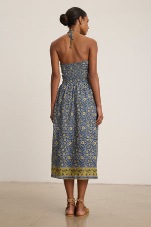 A woman faces away in the ODETTE DRESS by Velvet by Graham & Spencer—an ankle-length, chic halter dress with blue and yellow floral block print cotton—paired with flat sandals.