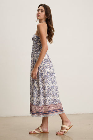 The woman stands in the ODETTE DRESS by Velvet by Graham & Spencer—a strapless, block print cotton midi dress with blue and red patterns—paired with beige sandals against a plain light background.