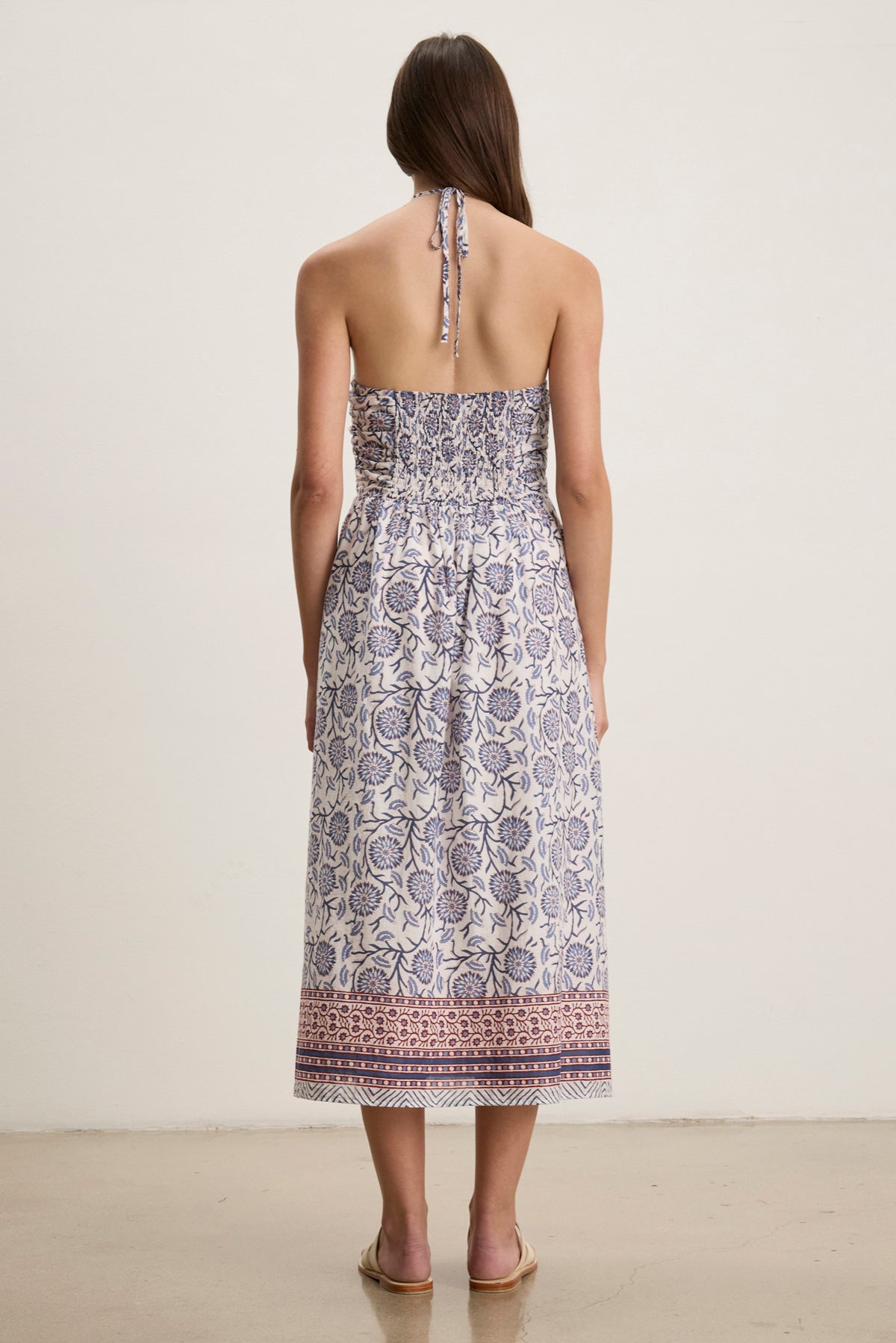 A woman faces away in the Velvet by Graham & Spencer ODETTE DRESS—a chic, ankle-length halter dress in blue and white floral block print cotton—paired with flat sandals.-39123350552769