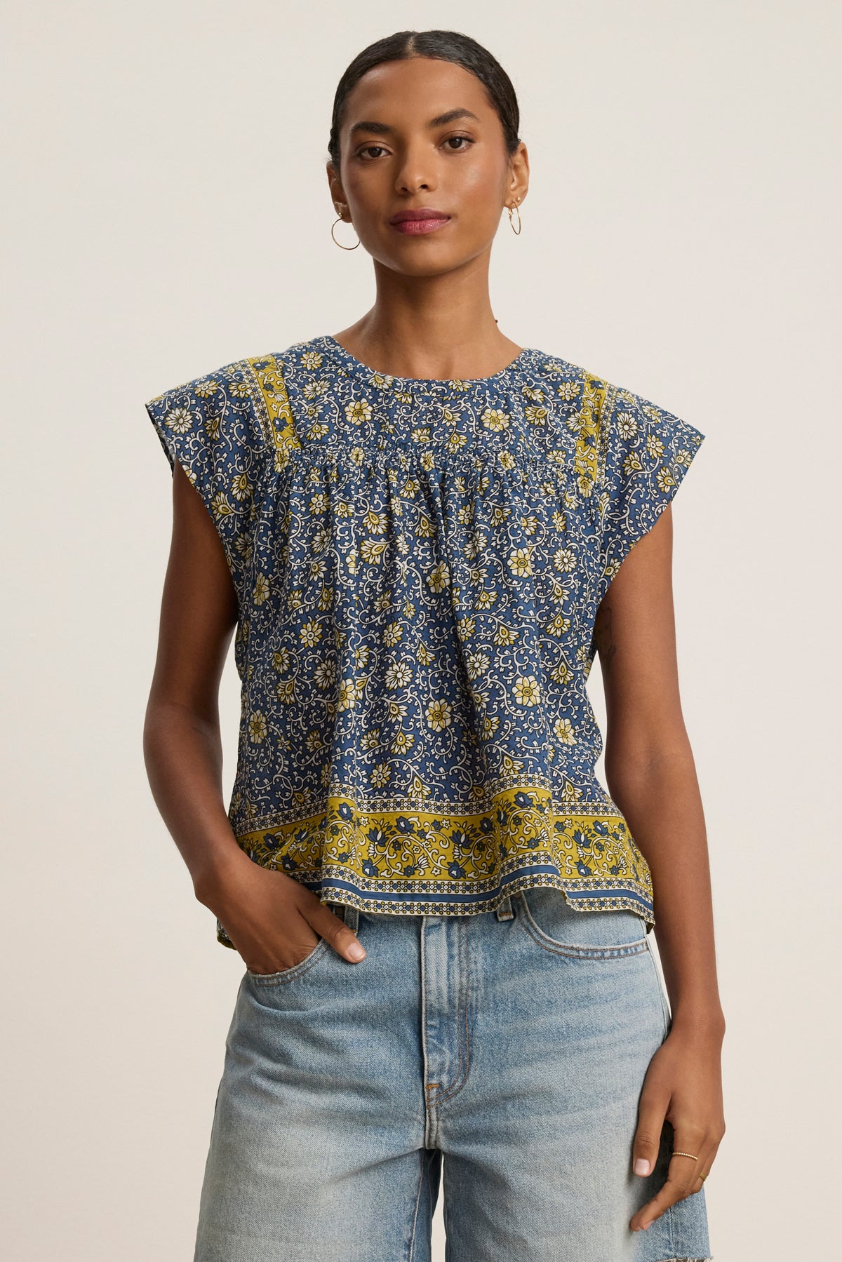 A woman models the SELENE TOP by Velvet by Graham & Spencer, a chic cropped top with blue and yellow block print florals and cap sleeves, paired with light blue jeans against a plain background.-39120162717889
