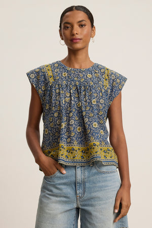 A woman models the SELENE TOP by Velvet by Graham & Spencer, a chic cropped top with blue and yellow block print florals and cap sleeves, paired with light blue jeans against a plain background.
