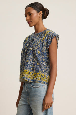 A woman wears the SELENE TOP by Velvet by Graham & Spencer, a chic blue and yellow block print sleeveless blouse, paired with light blue jeans against a plain light background.
