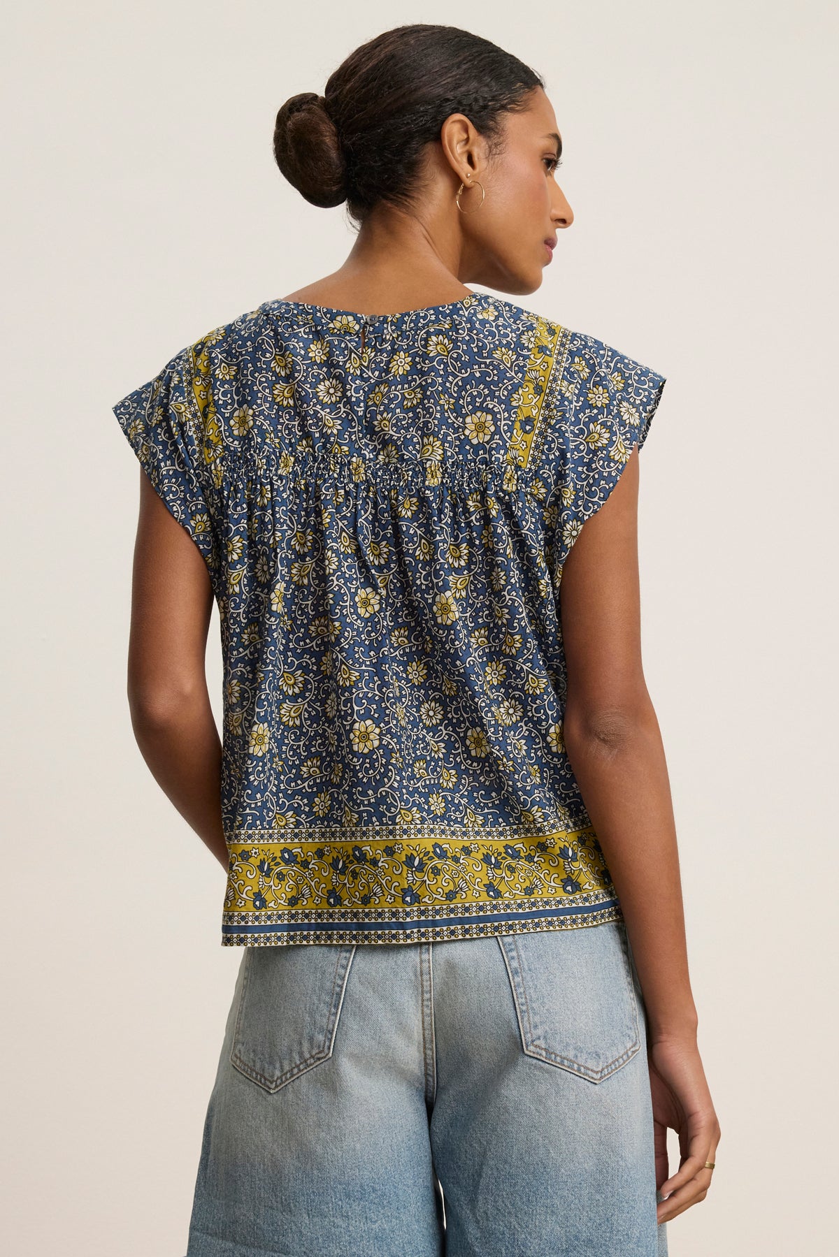 A woman with her hair in a bun wears the SELENE TOP by Velvet by Graham & Spencer—a chic, block print blue and yellow floral cropped blouse—paired with light blue jeans, shown from the back against a plain background.-39120162652353