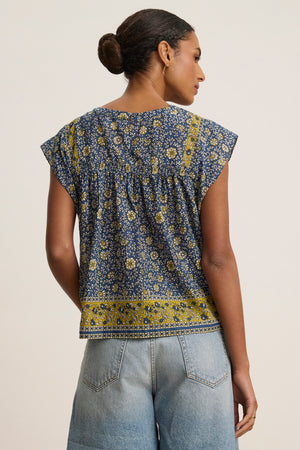 A woman with her hair in a bun wears the SELENE TOP by Velvet by Graham & Spencer—a chic, block print blue and yellow floral cropped blouse—paired with light blue jeans, shown from the back against a plain background.