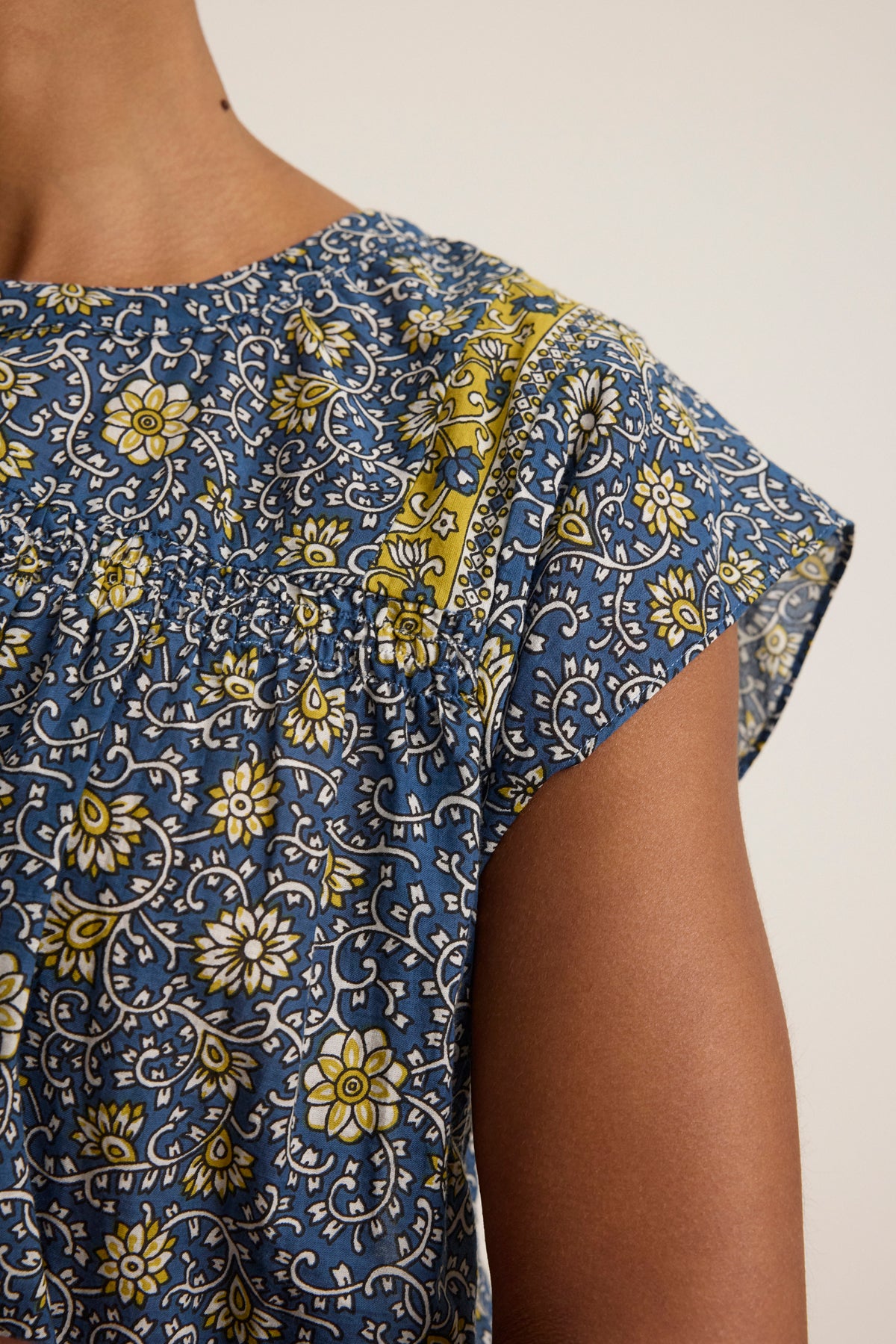 Close-up of a person wearing the SELENE TOP by Velvet by Graham & Spencer, a chic blue, short-sleeved cropped top with yellow and white floral block print and a decorative yoke detail.