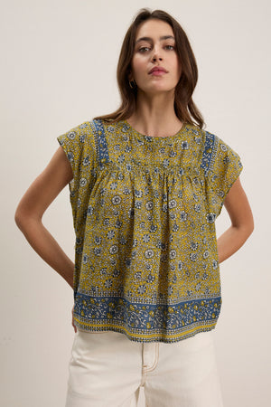 A woman wears the SELENE TOP by Velvet by Graham & Spencer—a yellow and blue floral block print top with short sleeves and a boxy cropped fit—paired with white pants, standing against a plain background.