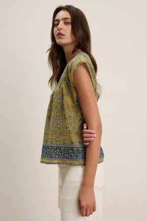 A woman stands sideways against a plain background, wearing the SELENE TOP by Velvet by Graham & Spencer—a light cotton, yellow and blue block print cropped top—paired with white pants.