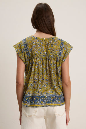 A woman with long brown hair, seen from behind, wears the Velvet by Graham & Spencer SELENE TOP— a chic yellow and blue block print short-sleeve top—paired with light-colored pants against a plain background.
