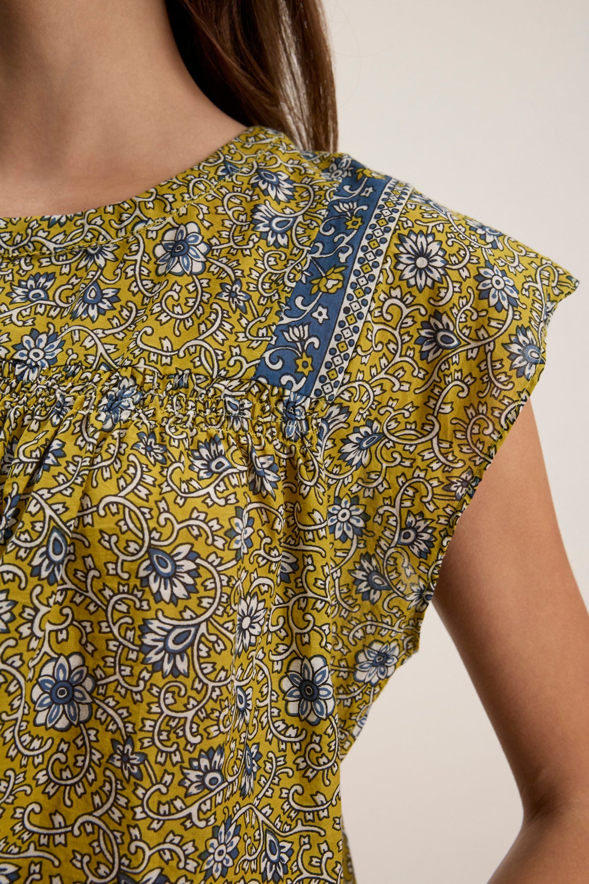 Close-up of a person wearing the SELENE TOP by Velvet by Graham & Spencer—a chic, cropped fit yellow floral-patterned top with blue and white accents and short sleeves.