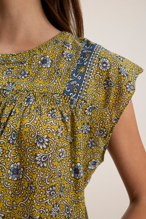 Close-up of a person wearing the SELENE TOP by Velvet by Graham & Spencer—a chic, cropped fit yellow floral-patterned top with blue and white accents and short sleeves.