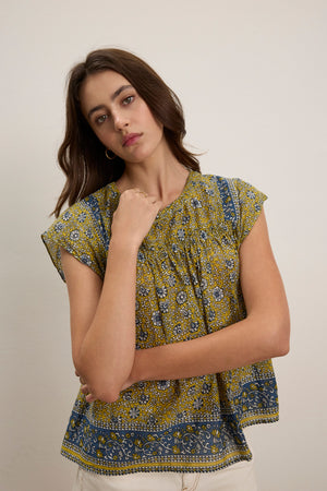 A woman with long brown hair wears the SELENE TOP by Velvet by Graham & Spencer—a chic yellow and blue block print blouse with short sleeves and a cropped fit—standing against a plain, light-colored background.