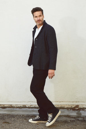 A man in a Velvet by Graham & Spencer Bowen Boiled Wool Blend Jacket and sneakers leaning against a wall.