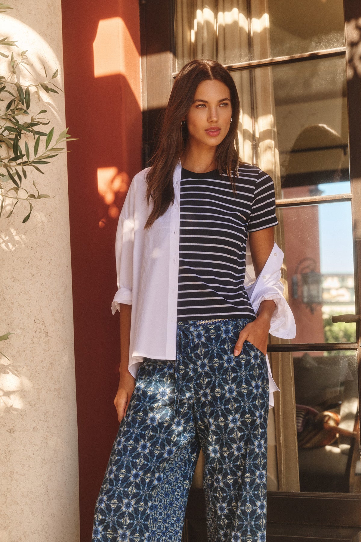 Outdoors, a woman wears the Velvet by Graham & Spencer BRENDA TEE—a navy striped cropped top with a fitted silhouette—layered over a white button-down shirt and paired with blue patterned wide-leg pants, one hand in her pocket.-40959850479809