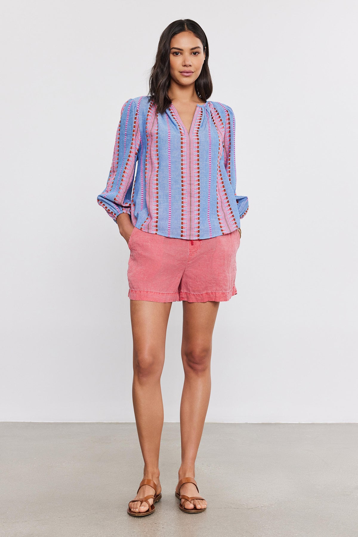 A woman stands wearing a Velvet by Graham & Spencer ISLA TOP with a v-neckline, coral shorts, and brown sandals, posing for the camera in a studio setting.-36910083997889