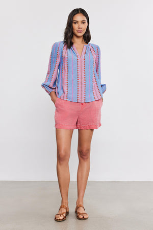 A woman stands wearing a Velvet by Graham & Spencer ISLA TOP with a v-neckline, coral shorts, and brown sandals, posing for the camera in a studio setting.