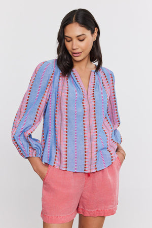 A woman in an ISLA TOP from Velvet by Graham & Spencer, with a v-neckline and coral shorts, standing against a white background.