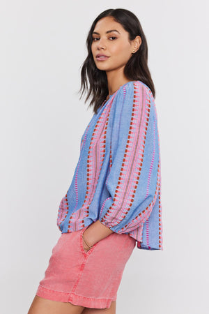 A woman in profile wearing a blue Jacquard stripe fabric ISLA TOP by Velvet by Graham & Spencer and pink shorts, looking over her shoulder.